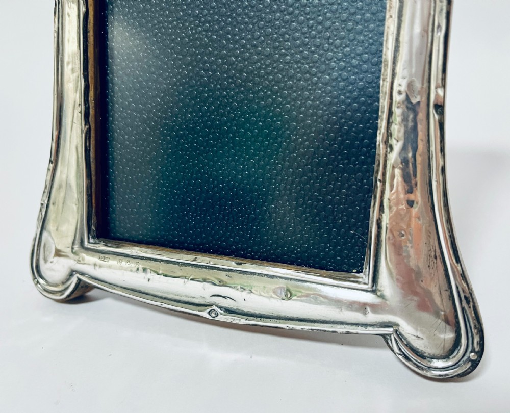 An antique silver picture frame featuring a curved scalloped rim, visible hallmarks along the lower border, and a dark textured interior display panel. View VIII