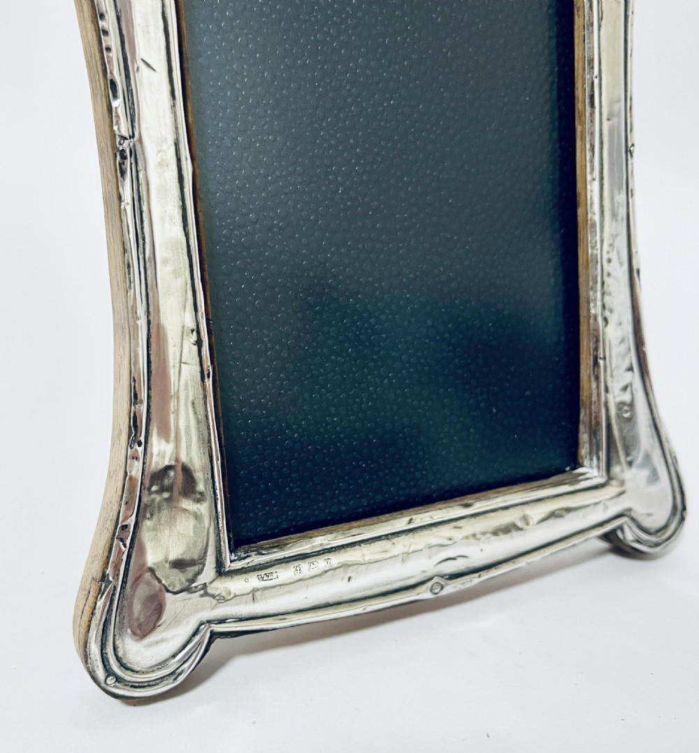 An antique silver picture frame featuring a curved scalloped rim, visible hallmarks along the lower border, and a dark textured interior display panel. View X