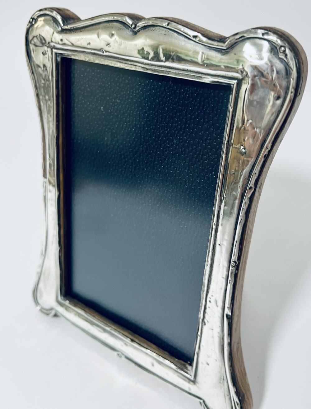 An antique silver picture frame featuring a curved scalloped rim, visible hallmarks along the lower border, and a dark textured interior display panel. View XI