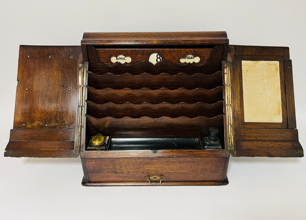 An open Victorian dark oak stationery cabinet featuring fitted letter slots, an integrated perpetual calendar, twin inkwells, and hinged side doors.