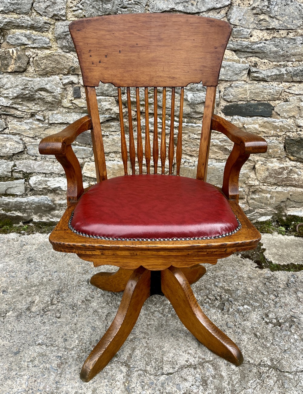 An Edwardian oak swivel desk chair featuring a spindle backrest, curved armrests, a scalloped apron, and a padded red leather seat trimmed with studs.