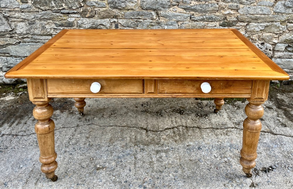 A large Victorian pine farmhouse table featuring a planked top, two frieze drawers with white ceramic knobs, and turned legs fitted with brass castors.