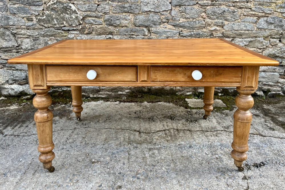 A large Victorian pine farmhouse table featuring a planked top, two frieze drawers with white ceramic knobs, and turned legs fitted with brass castors. View II
