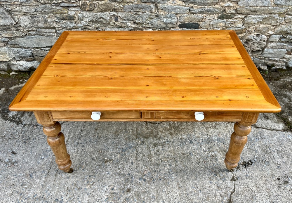 A large Victorian pine farmhouse table featuring a planked top, two frieze drawers with white ceramic knobs, and turned legs fitted with brass castors. View III