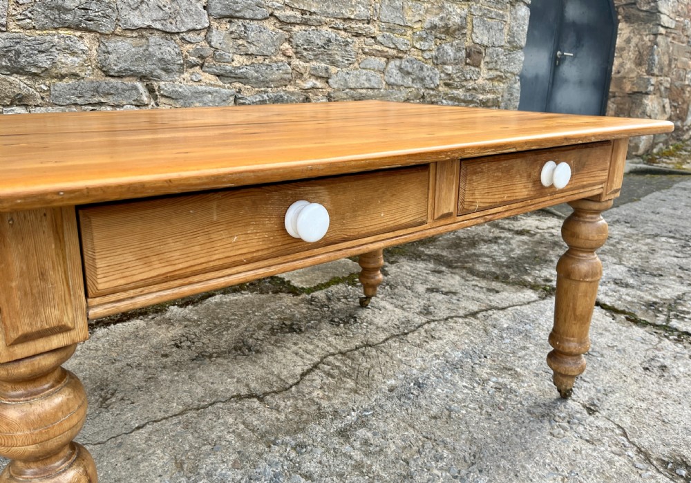 A large Victorian pine farmhouse table featuring a planked top, two frieze drawers with white ceramic knobs, and turned legs fitted with brass castors. View V