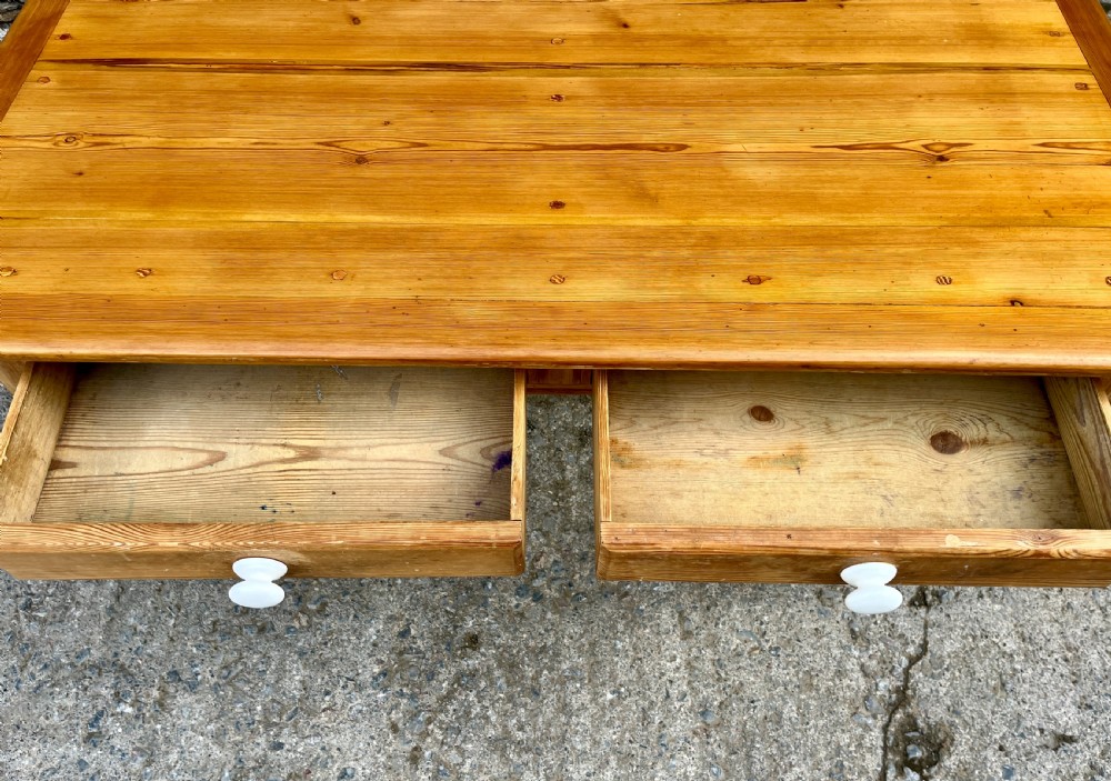 A large Victorian pine farmhouse table featuring a planked top, two frieze drawers with white ceramic knobs, and turned legs fitted with brass castors. View VI