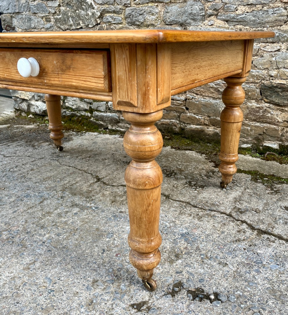 A large Victorian pine farmhouse table featuring a planked top, two frieze drawers with white ceramic knobs, and turned legs fitted with brass castors. View VII