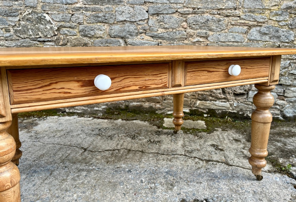 A large Victorian pine farmhouse table featuring a planked top, two frieze drawers with white ceramic knobs, and turned legs fitted with brass castors. View VIII