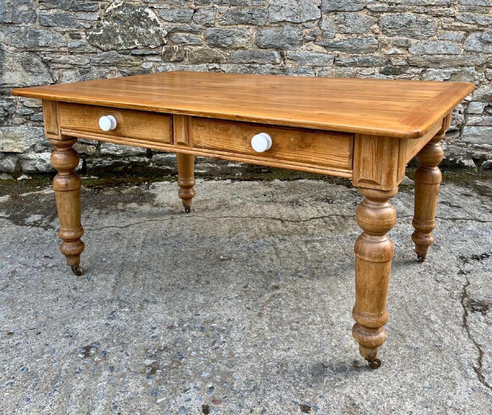 A large Victorian pine farmhouse table featuring a planked top, two frieze drawers with white ceramic knobs, and turned legs fitted with brass castors. View IX
