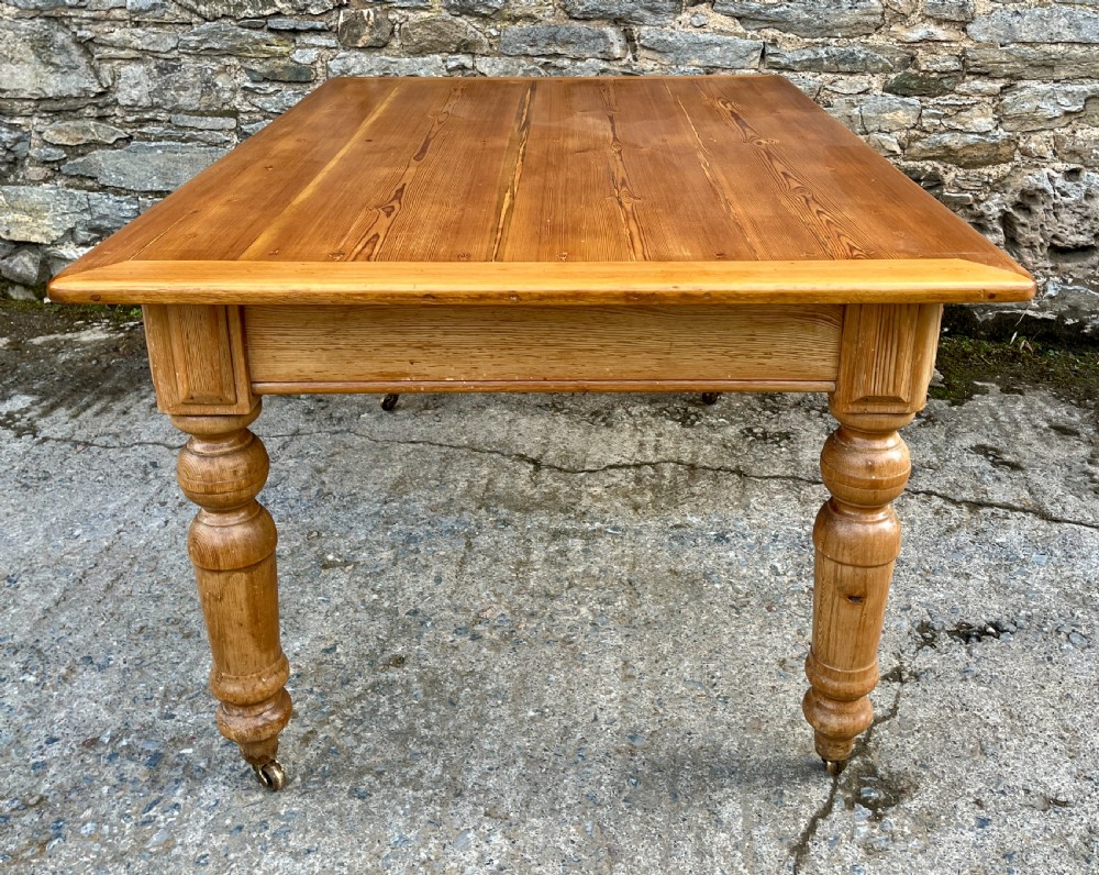 A large Victorian pine farmhouse table featuring a planked top, two frieze drawers with white ceramic knobs, and turned legs fitted with brass castors. View X