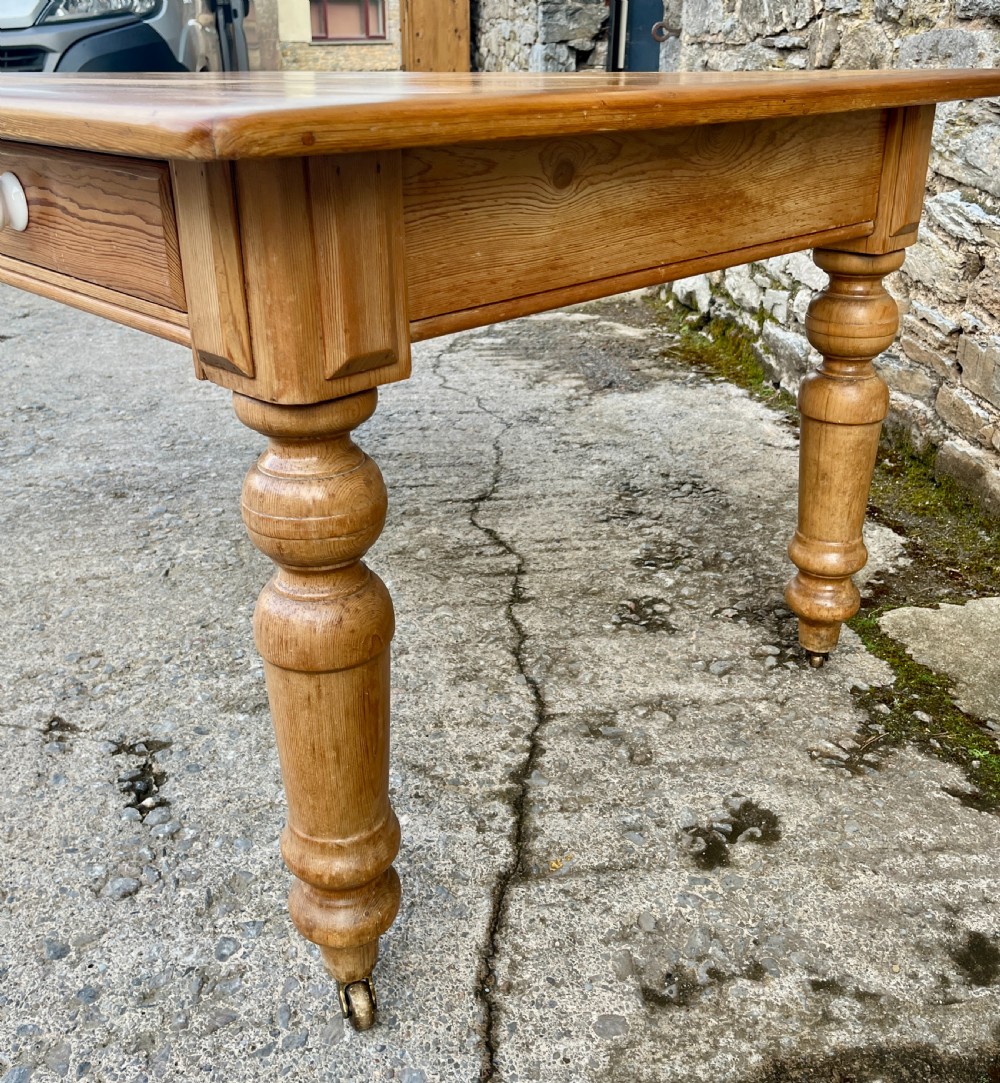 A large Victorian pine farmhouse table featuring a planked top, two frieze drawers with white ceramic knobs, and turned legs fitted with brass castors. View XI