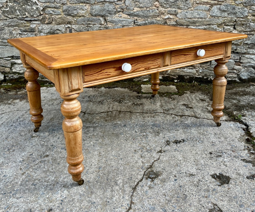 A large Victorian pine farmhouse table featuring a planked top, two frieze drawers with white ceramic knobs, and turned legs fitted with brass castors. View XIII