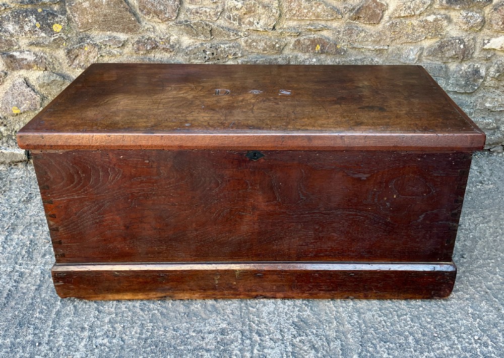 A rectangular dark teak blanket chest featuring carved initials on the hinged lid, exposed dovetail joints, and a keyhole escutcheon on the front.