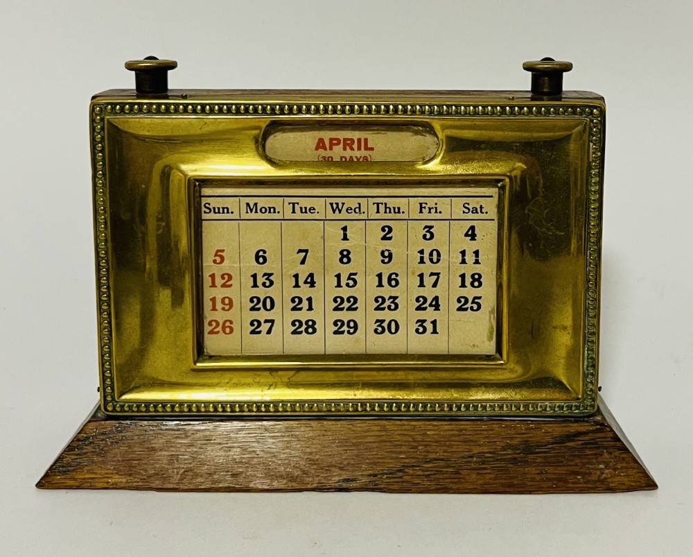 An antique brass perpetual desk calendar with a beaded frame mounted on a dark oak wooden base.
