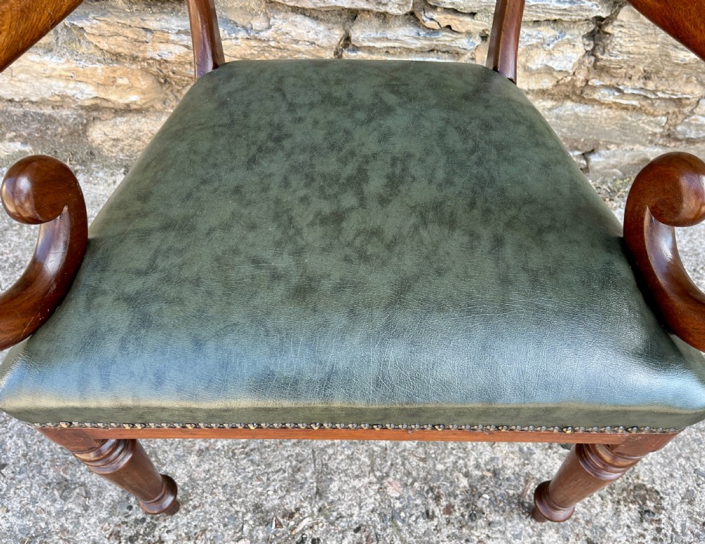 Victorian mahogany carver chair featuring dramatically scrolled armrests, a curved top rail, turned front legs, and a dark green leather seat trimmed with brass studs. View V