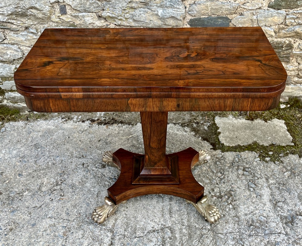 An antique Regency rosewood card table featuring a rectangular fold-over top, central tapered pedestal support, and a quadform base with brass claw feet. View II