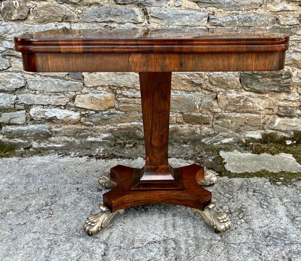 An antique Regency rosewood card table featuring a rectangular fold-over top, central tapered pedestal support, and a quadform base with brass claw feet. View III