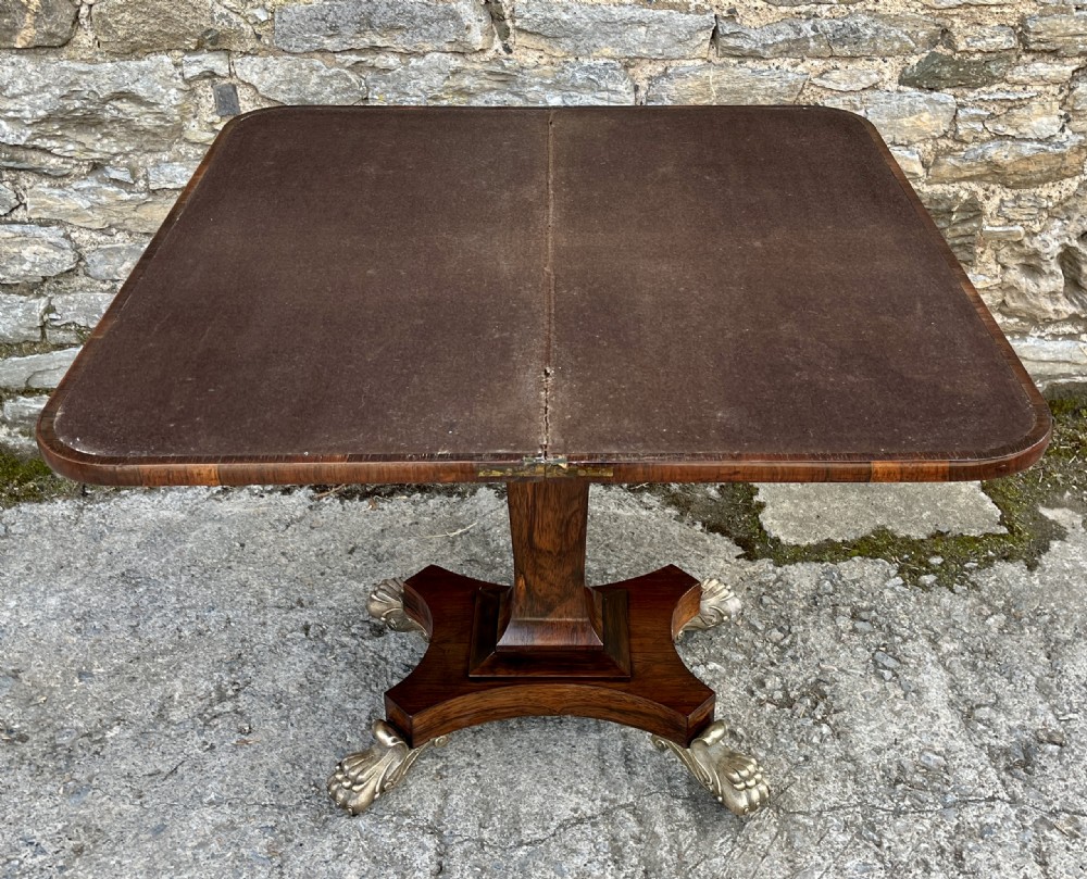 An antique Regency rosewood card table featuring a rectangular fold-over top, central tapered pedestal support, and a quadform base with brass claw feet. View X