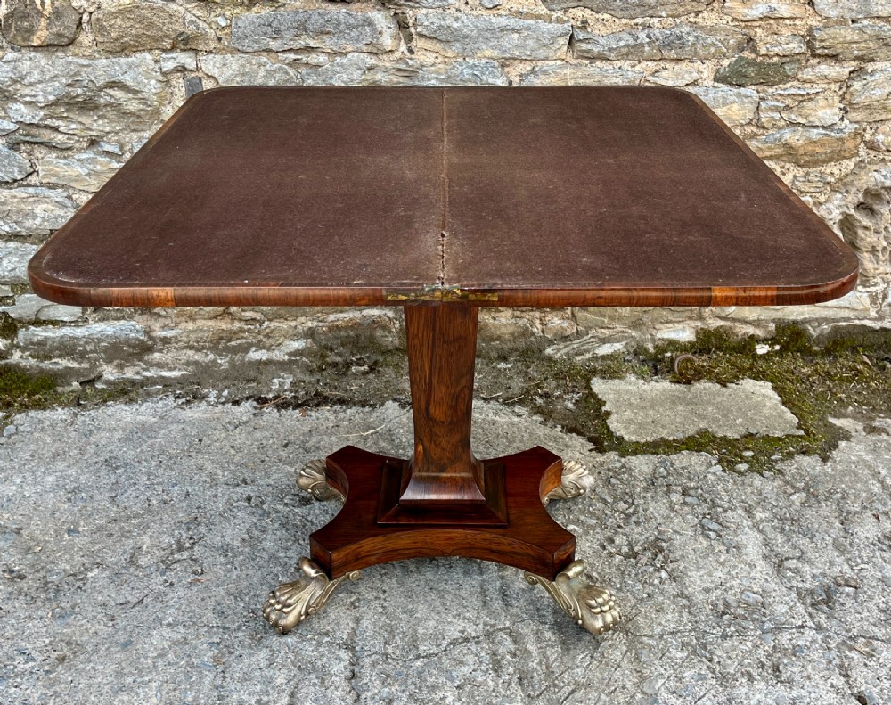 An antique Regency rosewood card table featuring a rectangular fold-over top, central tapered pedestal support, and a quadform base with brass claw feet. View XI
