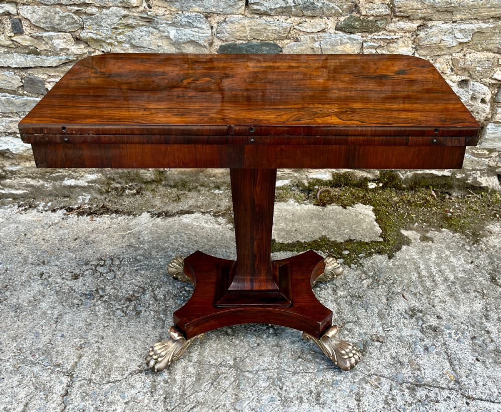 An antique Regency rosewood card table featuring a rectangular fold-over top, central tapered pedestal support, and a quadform base with brass claw feet. View XVII