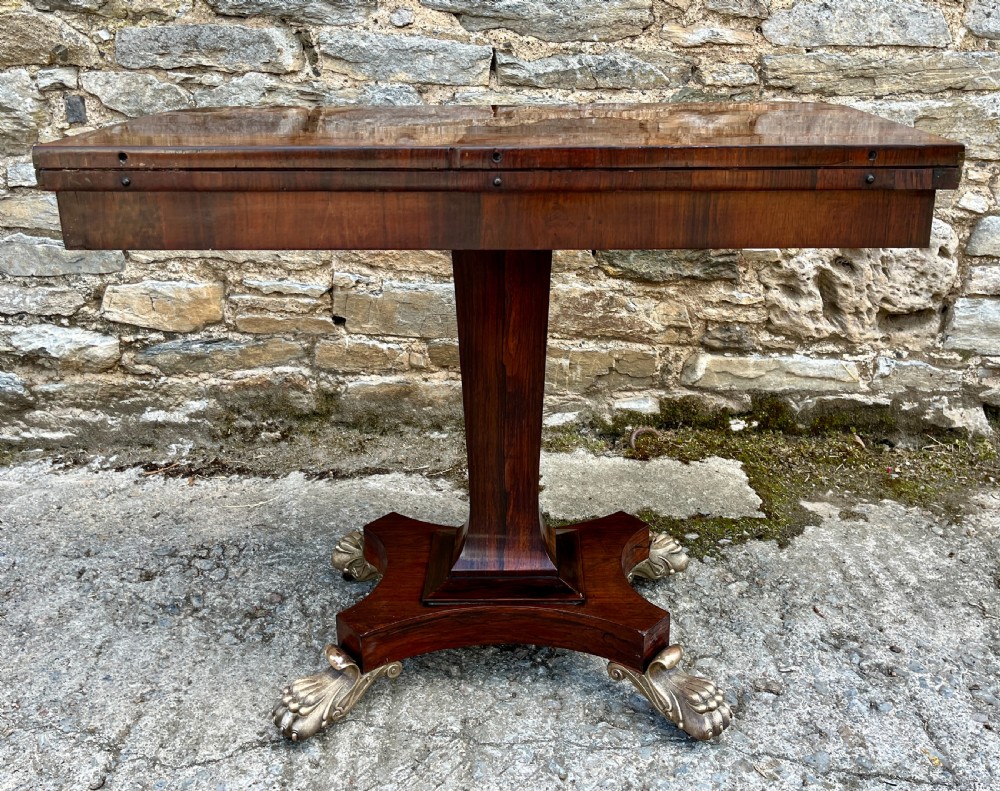 An antique Regency rosewood card table featuring a rectangular fold-over top, central tapered pedestal support, and a quadform base with brass claw feet. View XVIII