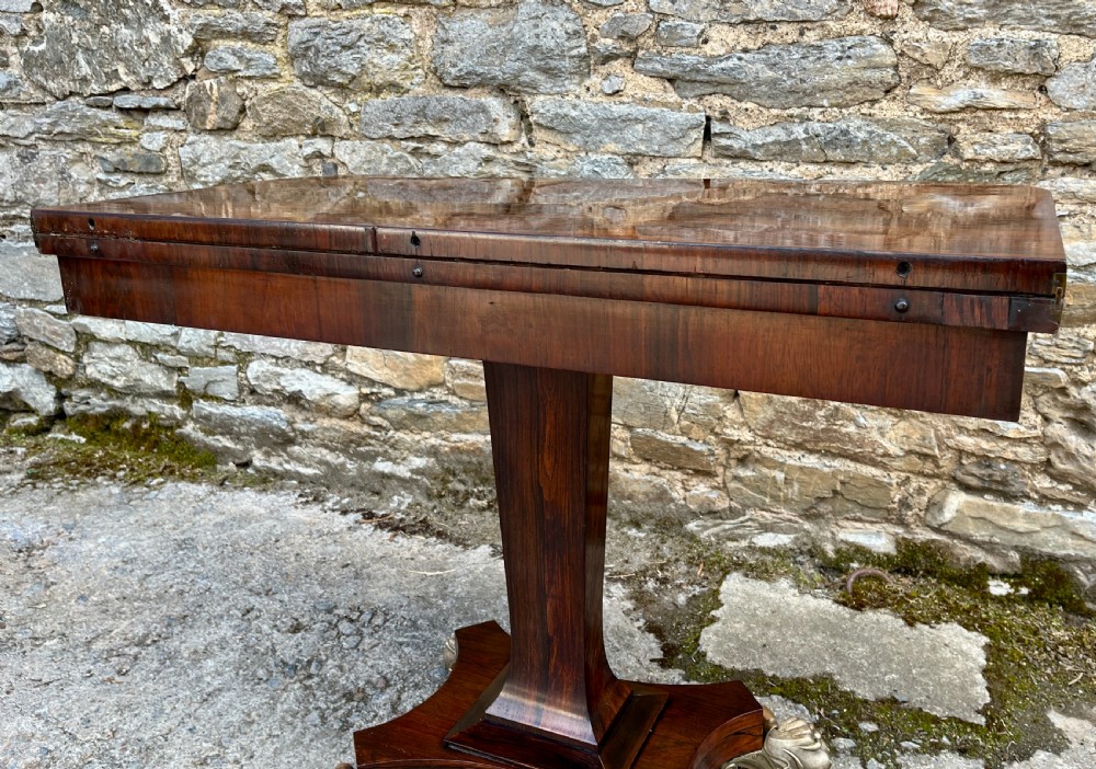 An antique Regency rosewood card table featuring a rectangular fold-over top, central tapered pedestal support, and a quadform base with brass claw feet. View XXI