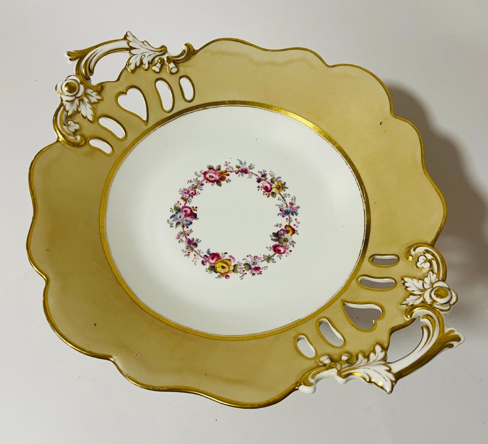 A nineteenth-century John Ridgway porcelain dessert service featuring buff-coloured borders, gilded scrollwork, reticulated handles, and hand-painted central floral garlands. View XIII