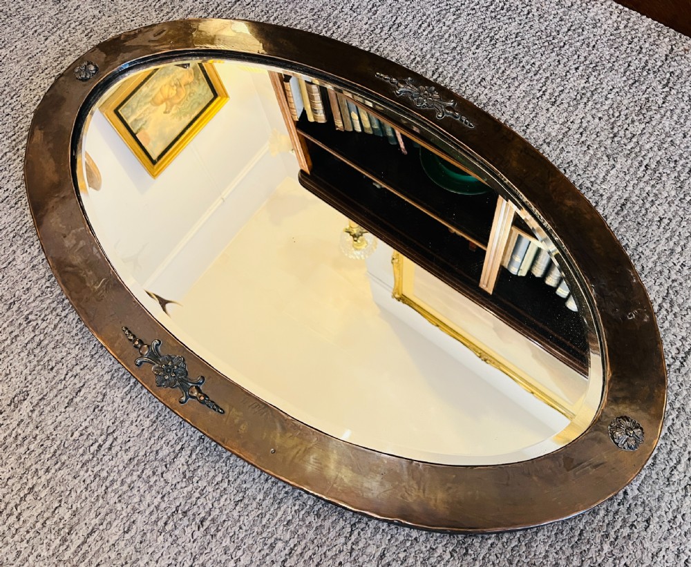 An oval Arts and Crafts wall mirror with a hand-hammered copper frame featuring embossed floral motifs at four points and a bevelled glass edge. View III