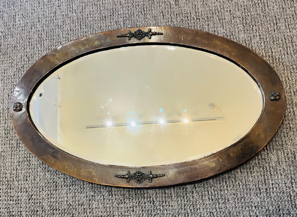 An oval Arts and Crafts wall mirror with a hand-hammered copper frame featuring embossed floral motifs at four points and a bevelled glass edge. View VII