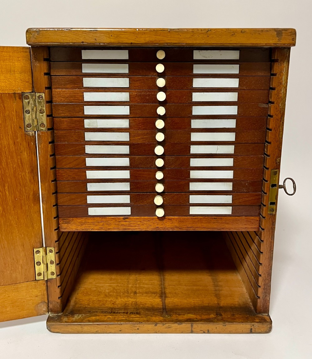 A warm brown wooden microscope slide cabinet with its single door open to reveal numerous shallow horizontal drawers with small round pulls. View II