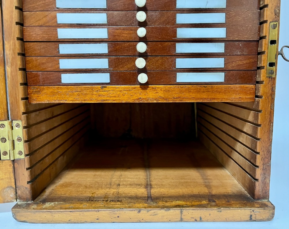 A warm brown wooden microscope slide cabinet with its single door open to reveal numerous shallow horizontal drawers with small round pulls. View V
