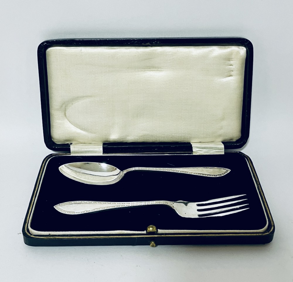 An antique silver christening spoon and fork set presented inside an open black fitted case lined with cream silk and dark velvet.