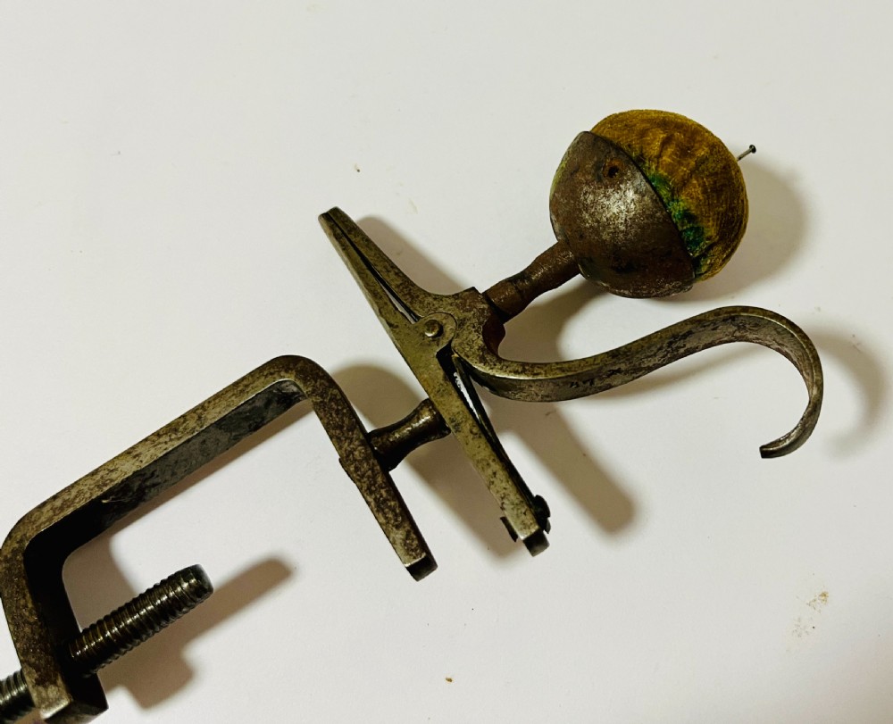 A Victorian dark steel table-mounted sewing clamp featuring a threaded screw, spring mechanism, curved hook, and a spherical fabric pincushion at the top. View II