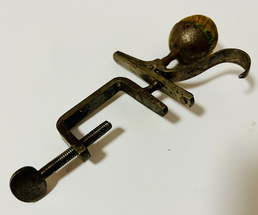 A Victorian dark steel table-mounted sewing clamp featuring a threaded screw, spring mechanism, curved hook, and a spherical fabric pincushion at the top. View III