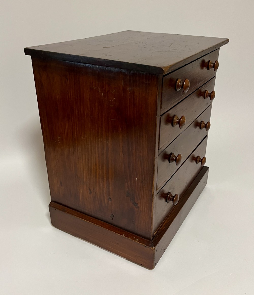 A small antique stained pine tabletop collectors cabinet featuring four drawers with turned wooden knobs on a plinth base. View X