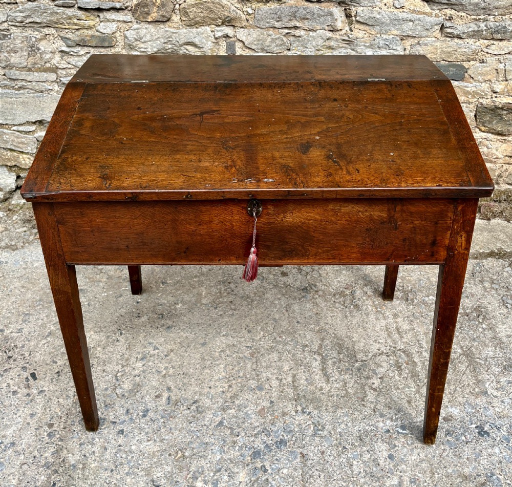 A Georgian solid oak clerks desk featuring a hinged sloped writing top, a key with tassel inserted into the central keyhole, and four square tapered legs.