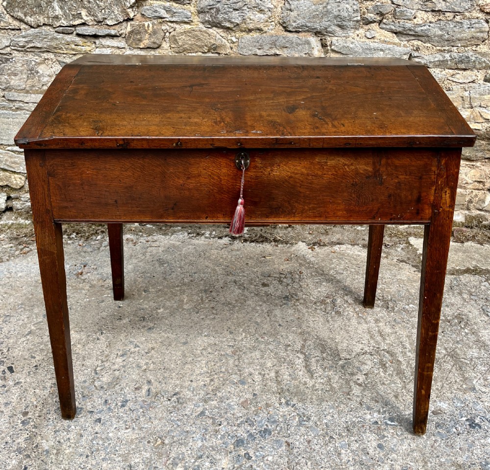 A Georgian solid oak clerks desk featuring a hinged sloped writing top, a key with tassel inserted into the central keyhole, and four square tapered legs. View II