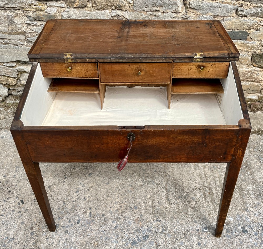 A Georgian solid oak clerks desk featuring a hinged sloped writing top, a key with tassel inserted into the central keyhole, and four square tapered legs. View III