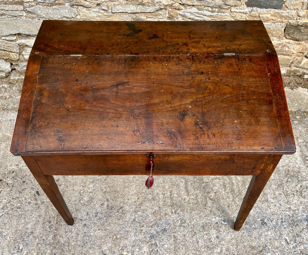 A Georgian solid oak clerks desk featuring a hinged sloped writing top, a key with tassel inserted into the central keyhole, and four square tapered legs. View IV