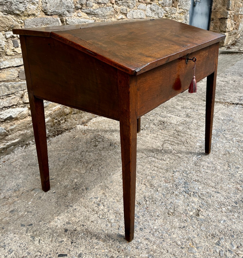 A Georgian solid oak clerks desk featuring a hinged sloped writing top, a key with tassel inserted into the central keyhole, and four square tapered legs. View V