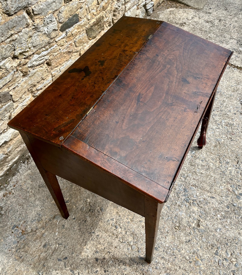 A Georgian solid oak clerks desk featuring a hinged sloped writing top, a key with tassel inserted into the central keyhole, and four square tapered legs. View VI