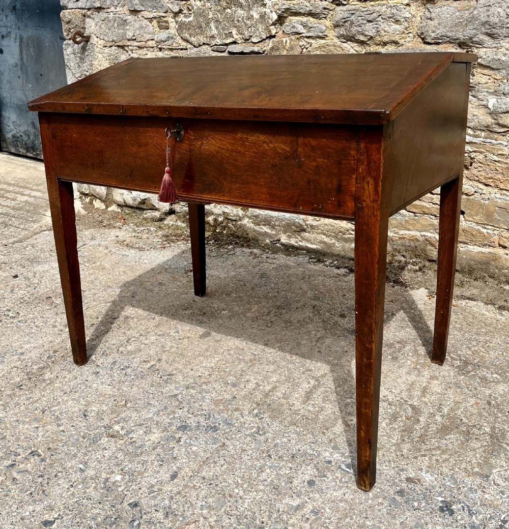 A Georgian solid oak clerks desk featuring a hinged sloped writing top, a key with tassel inserted into the central keyhole, and four square tapered legs. View VII