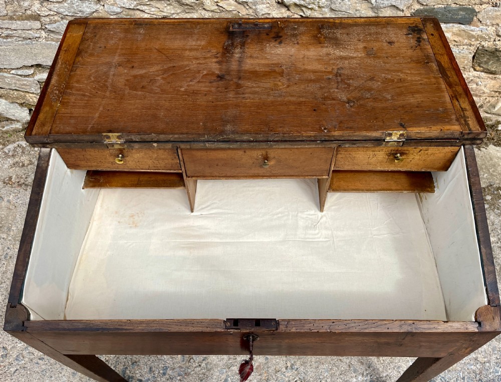 A Georgian solid oak clerks desk featuring a hinged sloped writing top, a key with tassel inserted into the central keyhole, and four square tapered legs. View IX