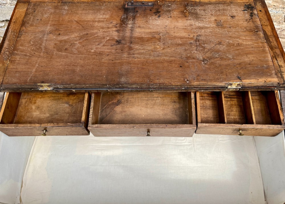 A Georgian solid oak clerks desk featuring a hinged sloped writing top, a key with tassel inserted into the central keyhole, and four square tapered legs. View X