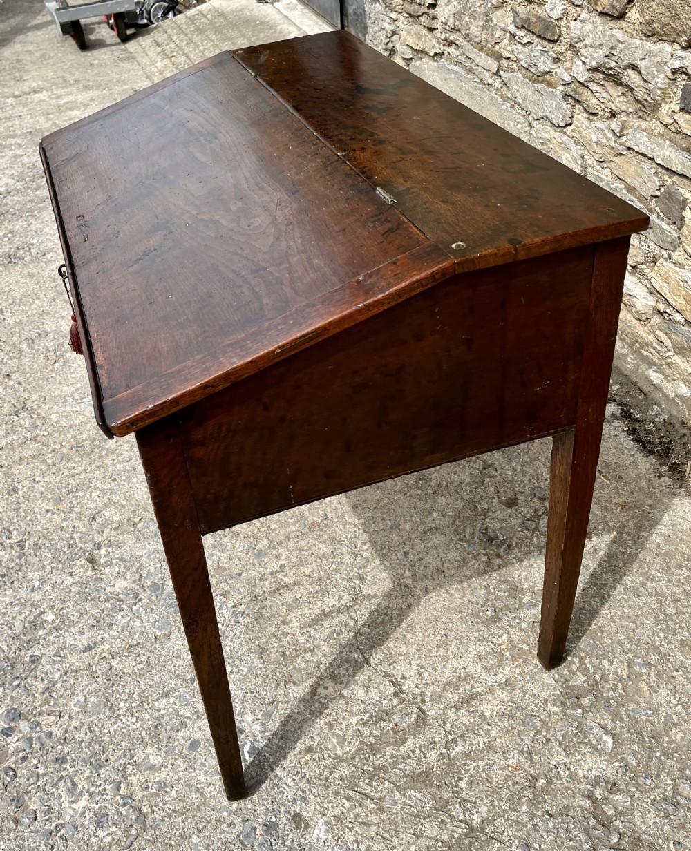 A Georgian solid oak clerks desk featuring a hinged sloped writing top, a key with tassel inserted into the central keyhole, and four square tapered legs. View XIII