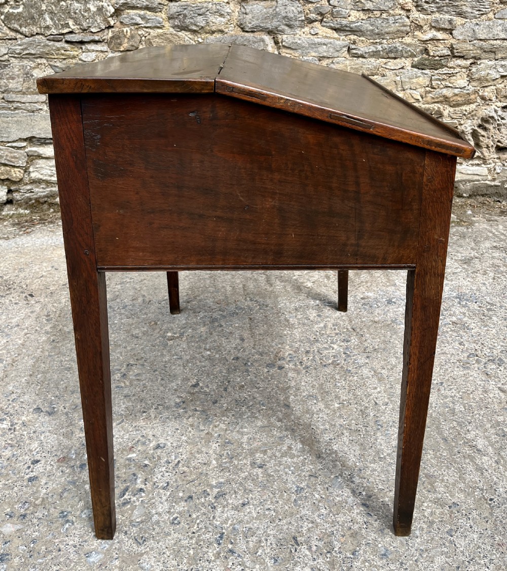 A Georgian solid oak clerks desk featuring a hinged sloped writing top, a key with tassel inserted into the central keyhole, and four square tapered legs. View XVII