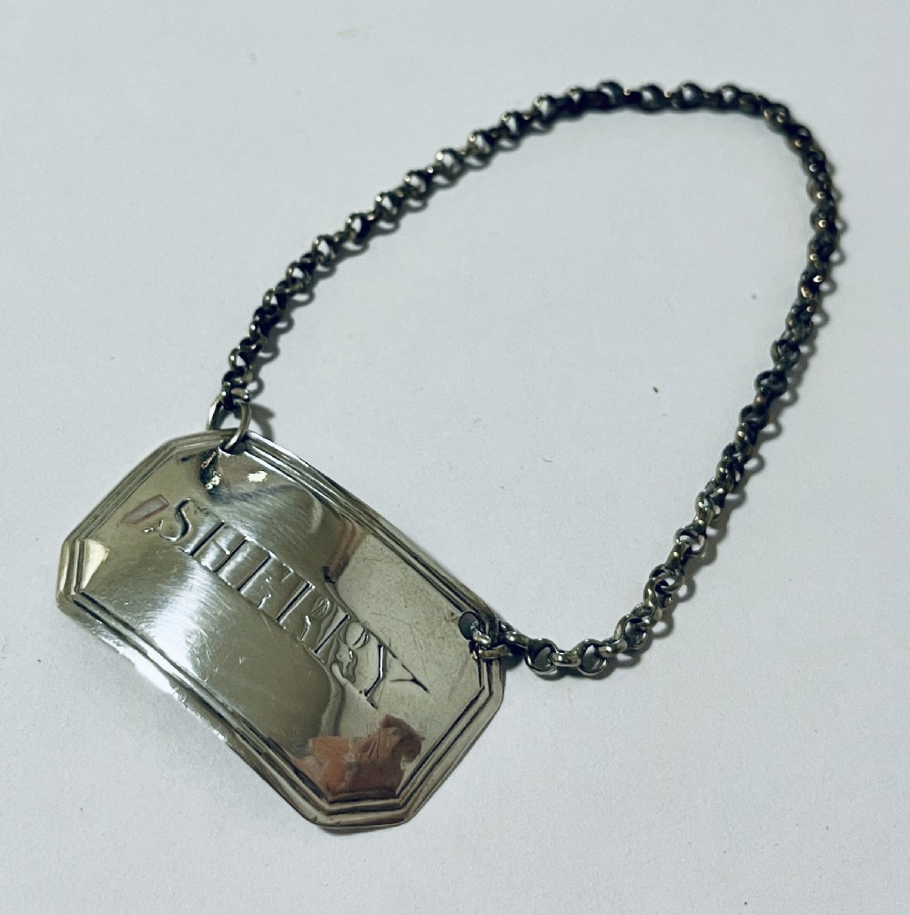 A rectangular silver decanter label with chamfered corners, engraved with the word SHERRY and suspended from a fine silver chain.