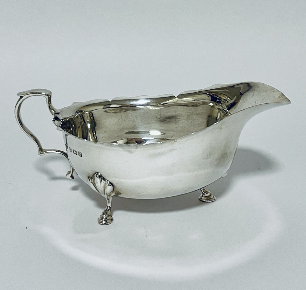 A polished sterling silver sauce boat featuring a cut rim, flying scroll handle, shell-capped tripod feet, and visible side hallmarks.