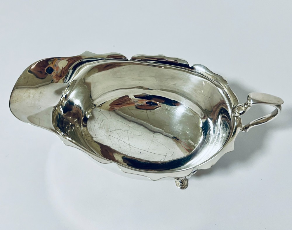A polished sterling silver sauce boat featuring a cut rim, flying scroll handle, shell-capped tripod feet, and visible side hallmarks. View VII