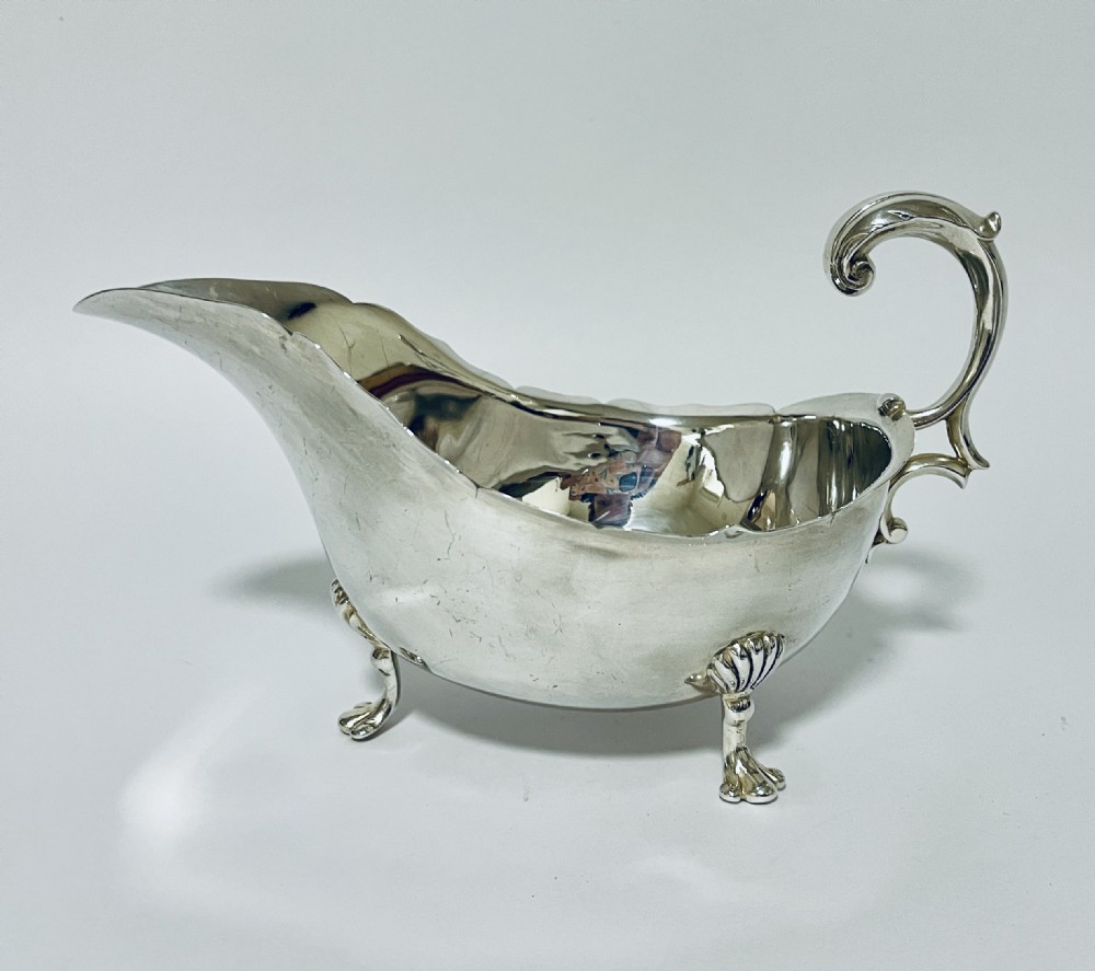 A polished antique silver sauce boat featuring a wavy scalloped rim, a double scroll handle, and three shell-capped cabriole feet.
